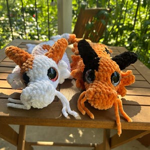 Puede incluir: Dos juguetes de dragón de peluche tejidos a mano. El dragón de la izquierda es blanco con manchas naranjas y ojos oscuros grandes. El dragón de la derecha es naranja con manchas negras, cuernos negros y ojos oscuros grandes. Ambos tienen bigotes.