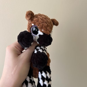 May include: A plush okapi toy crafted from soft brown, black, and white yarn. The stuffed animal features a textured, fuzzy appearance, large dark purple glittery eyes, and the distinct black and white striped leg markings characteristic of an okapi.
