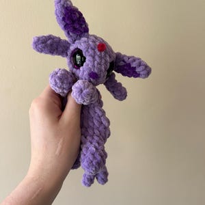 Puede incluir: Un peluche de ganchillo hecho a mano con hilo morado suave. La criatura presenta orejas largas con detalles en morado oscuro, grandes ojos negros brillantes, un pequeño punto rojo en la frente y un punto morado en cada mejilla, con cuerpo.