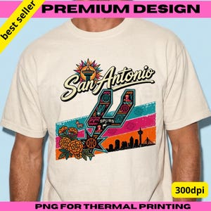 May include: A cream-colored t-shirt featuring a graphic design with the text San Antonio. The design includes a stylized spur logo, orange marigold flowers, a piñata, a city skyline silhouette, and colorful diagonal stripes in teal, pink, and orange.