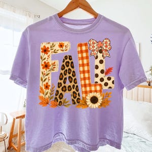 Peut inclure: Un t-shirt à manches courtes violet clair avec le mot FALL imprimé sur le devant. La lettre F présente un motif floral, le A un imprimé léopard, le L un motif vichy orange et le L des pois noirs avec un nœud décoratif au-dessus.