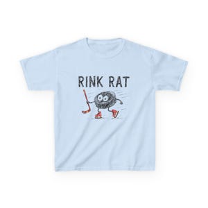 Puede incluir: Camiseta azul claro con el texto "RINK RAT" en fuente negra desgastada. El gráfico presenta un personaje de disco de hockey de dibujos animados con patines rojos y un palo de hockey. El disco tiene ojos grandes y está sonriendo.