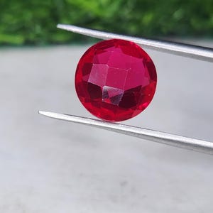 May include: A round, faceted, vibrant red gemstone held by a pair of silver-toned metal tweezers. The stone features a brilliant cut with multiple geometric facets that reflect light, showcasing a deep, saturated crimson color throughout the surface.