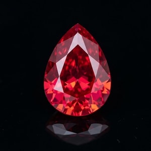 Puede incluir: Una piedra preciosa en forma de pera de color rojo vibrante con una superficie facetada que crea reflejos de luz geométricos. La piedra está colocada sobre un fondo negro sólido, mostrando un reflejo claro de su forma puntiaguda en la base.