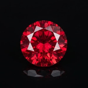 May include: A round, brilliant-cut gemstone with a deep, vibrant red color. The stone features numerous triangular and kite-shaped facets that create a complex, symmetrical pattern of light reflection and refraction across its surface.