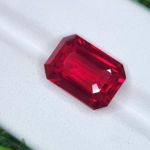 May include: A rectangular-cut ruby gemstone with multiple facets. The gem is a deep red colour and is set against a white surface. The facets reflect light, creating a sparkling effect. This image showcases a precious stone.