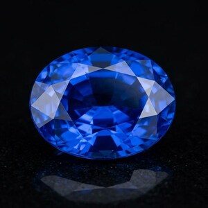 Puede incluir: Una gema azul de corte ovalado con una superficie facetada que refleja la luz. La piedra muestra un color azul real profundo y saturado con patrones geométricos claros creados por el corte de precisión sobre un fondo oscuro.