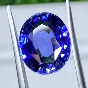 May include: A deep blue, oval-cut gemstone is held by silver tweezers. The gem's facets reflect light, creating a sparkling effect. The colour is a rich, saturated blue, with variations in tone across the surface. The gem is likely a sapphire.