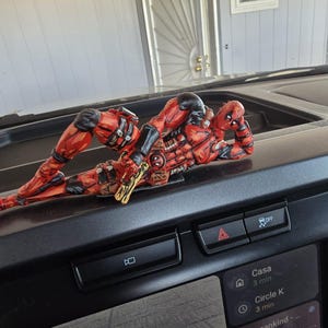 Könnte beinhalten: Eine rot-schwarze Deadpool-Figur mit einer goldfarbenen Pistole liegt auf dem Armaturenbrett eines Autos. Die Figur ist detailliert mit einer schwarzen Maske und einem roten Anzug. Das Armaturenbrett hat Knöpfe und einen Bildschirm mit einer Karte.