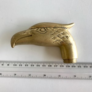 May include: A brass eagle head handle with a threaded base, positioned above a clear plastic ruler. The eagle head features engraved eye and feather details. The ruler shows the object measures approximately 13.3 cm in total length.