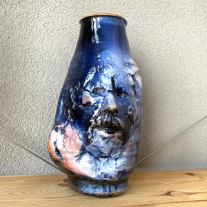 Könnte beinhalten: Eine hohe Keramikvase mit glänzender Oberfläche in den Farben Tiefblau, Weiß und kupferfarbenen Akzenten. Die Oberfläche zeigt ein skulpturales, dreidimensionales Relief eines menschlichen Gesichts mit detaillierten Zügen im Gefäßkörper.