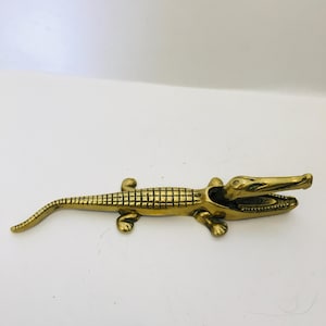 May include: A polished brass figurine of an alligator in a horizontal position. The alligator has a textured, grid-like pattern on its back, four legs, a long tail, and an open mouth revealing detailed teeth. The metal surface is gold-toned.