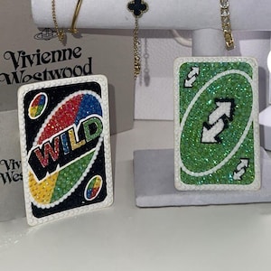 Puede incluir: Una variedad de joyas y objetos decorativos. Dos cartas de Uno adornadas, una con "WILD" en pedrería colorida, la otra con un fondo verde y un símbolo de inversión. También se exhiben pulseras doradas y un collar de trébol negro.