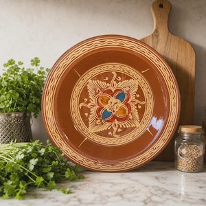 Può includere: Un piatto in ceramica marrone con un motivo floreale centrale con accenti rossi, blu e gialli. Il piatto ha un bordo decorativo ed è esposto con un tagliere in legno ed erbe fresche.