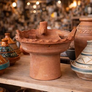 Puede incluir: Una olla de tagine de terracota con un borde festoneado decorativo y un asa central. La olla es de un color marrón rojizo cálido y se encuentra sobre una superficie de madera, rodeada de otras piezas de cerámica.