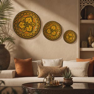 May include: Three decorative wall plates with a floral design in yellow, orange, and black. The plates are of varying sizes and are mounted on a neutral-colored wall. The plates are above a sofa with orange and cream-colored pillows.