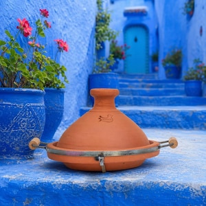 Puede incluir: Una olla de tagine de terracota con tapa cónica y banda metálica. La olla tiene asas de madera. El fondo presenta paredes azules, escaleras y plantas en macetas con flores rosas. La olla es de terracota.