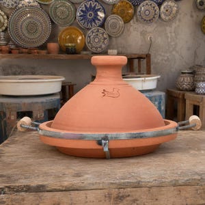 May include: A terracotta tagine pot with a conical lid and a wide, shallow base. The pot has a metal band around the base with wooden handles. The lid features a logo and a rounded top. The tagine is displayed on a wooden surface.