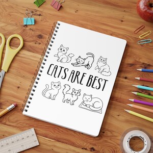 May include: A white spiral-bound notebook featuring the text "CATS ARE BEST" and cat illustrations. The notebook is on a wooden surface with stationery items, including pencils and scissors. The notebook is surrounded by stationery.