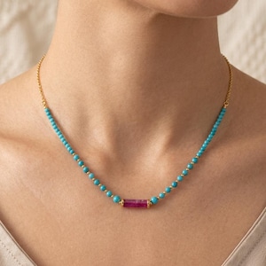 Puede incluir: Un collar delicado que presenta una serie de pequeñas cuentas redondas de color turquesa ensartadas en una cadena dorada, centrado con una cuenta de piedra preciosa magenta cilíndrica y alargada flanqueada por dos pequeños espaciadores dorados.