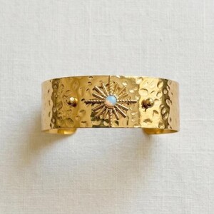 May include: A gold-toned cuff bracelet featuring a hammered, textured surface. The center of the band displays a prominent starburst design with a small, round, iridescent opal-like stone at its core, flanked by two smooth, raised gold spheres.