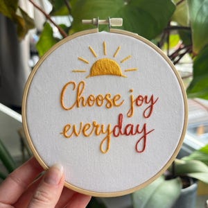 May include: Embroidered hoop art with the words "Choose joy everyday" in orange and red thread. A yellow sun design is above the text. The artwork is set in a wooden embroidery hoop.