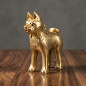 Könnte beinhalten: Eine goldfarbene Metallfigur eines stehenden Hundes mit spitzen Ohren und einem geringelten Schwanz. Die Skulptur weist eingravierte kreisförmige und lineare Muster um den Hals und entlang des Körpers auf, mit einer glatten, polierten Oberfläche.