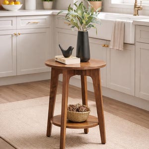 Puede incluir: Una mesa auxiliar redonda de madera con un estante inferior. Una jarrón negro con vegetación, una figura de pájaro negro y un libro pequeño están encima. Una cesta tejida con bolas decorativas está en el estante inferior.