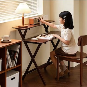May include: A dark brown wooden desk with a matching chair, a small wooden radio, and a lamp. A young person is sitting at the desk, reading a book. A cup and saucer are on a nearby shelf.