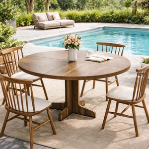 Pode incluir: Uma mesa de jantar redonda de madeira com quatro cadeiras a condizer. A mesa tem um acabamento em madeira escura e uma base de pedestal. As cadeiras têm almofadas de cor clara e um design de encosto de fuso. Um vaso de flores está sobre a mesa.