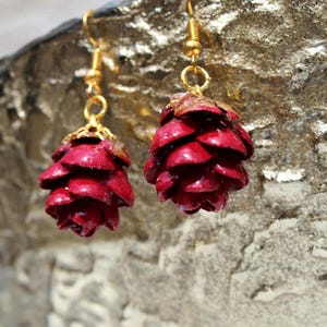 Peut inclure: Boucles d'oreilles pendantes représentant des pommes de pin. Les boucles d'oreilles sont d'un rouge profond, avec des crochets et des accents dorés. Les formes de pommes de pin sont détaillées et texturées, créant un aspect naturel.