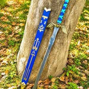 Könnte beinhalten: Ein Fantasy-Schwert mit silberner Klinge und goldenen Akzenten, das an einem Baum lehnt. Der Griff ist mit blauem und türkisfarbenem Leder umwickelt. Eine blaue Scheide mit goldenen Details liegt daneben, was auf einen thematischen Cosplay-Artikel hindeutet.