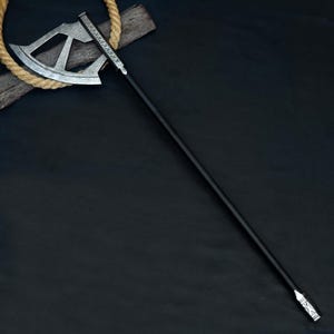May include: A silver and black battle axe with a long black handle. The axe head has a decorative design. The handle is long and slender. The axe is lying on a dark surface with a rope and wood in the background.