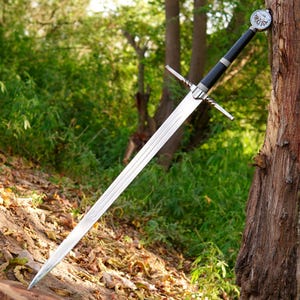 May include: A long steel sword with a double-edged blade, a silver crossguard, and a black wrapped handle. The pommel features a circular emblem with a winged crest design. The sword is positioned diagonally against a textured tree trunk.