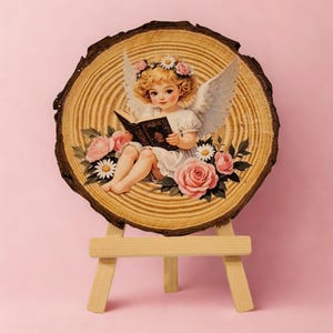 May include: A wooden slice featuring a vintage illustration of a cherubic figure with wings, reading a book, surrounded by pink roses and daisies. The artwork is displayed on a small wooden easel against a pink backdrop.