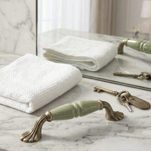 May include: A sage green ceramic drawer pull with an antique brass finish on the mounting ends, positioned near a white folded textured towel and a metal key with a tag labelled HOME, all resting on a white marble surface with grey veining.