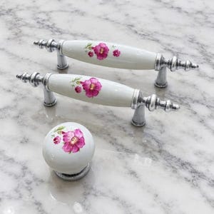 May include: Two white ceramic drawer pulls with a spindle shape and one matching round ceramic knob. Each piece features a floral pattern with bright pink flowers and green leaves, accented by polished silver-tone metal hardware.