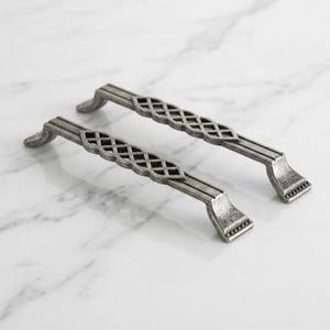 May include: Two silver-toned metal drawer pulls featuring a textured, weathered finish. Each handle has a central section with an open, crisscross lattice pattern and flared ends with small, horizontal grooved detailing on the mounting points.