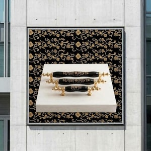 Può includere: Tre maniglie per armadietti nere con motivi floreali dorati ed estremità in metallo color oro, disposte a gradini su un blocco rettangolare bianco. Le maniglie presentano un centro cilindrico con un motivo ripetitivo di viti e fiori.