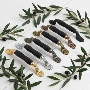 May include: A collection of five cabinet handles, each featuring a matte black central bar and metallic ends in gold, silver, gunmetal, brass, and copper tones. The handles have a curved, fluted design at the mounting points and a smooth grip.