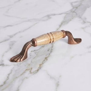 May include: A cabinet handle featuring a central cream-coloured ceramic barrel with a subtle speckled texture, flanked by two ornate, fluted metal ends finished in a weathered, antique copper tone with visible dark brown patina accents.