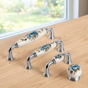 May include: A collection of three cabinet pulls and one matching round cabinet knob. Each piece features a white ceramic center with a blue floral pattern and brown leaves, accented by polished silver-tone metal hardware at the ends.