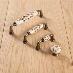 May include: A collection of four cabinet hardware pieces including three arched drawer pulls and one round knob. Each piece features a white ceramic center with a painted orange floral pattern and green leaves, finished with antique brass metal ends.