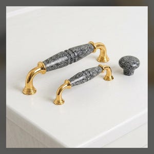 May include: Two gold-toned metal drawer pulls and one matching round knob. Each piece features a central section made of grey and black speckled stone-like material with polished gold-toned metal ends and mounting bases for cabinet hardware.