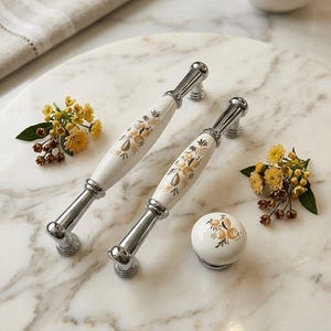 May include: Two elongated cabinet pulls and one round cabinet knob, each featuring a white ceramic center with a gold and brown floral pattern and polished silver-tone metal ends. The hardware is arranged on a white marble surface.