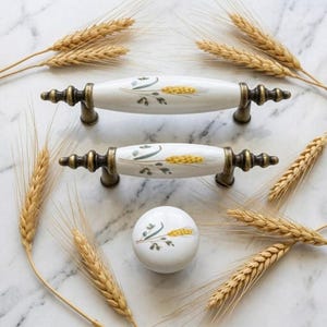 May include: Two white ceramic drawer pulls with an elongated oval shape and one round ceramic knob, all featuring a painted wheat stalk design and antique brass-toned metal hardware. The hardware is arranged with wheat stalks.