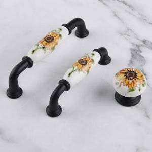 May include: Two cabinet pulls and one round cabinet knob featuring a white ceramic center with a painted yellow sunflower design and green leaves, finished with matte black metal hardware at the ends and base for mounting.