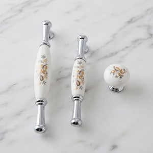 May include: Two elongated cabinet pulls and one round cabinet knob, all featuring a white ceramic center with a gold and grey floral pattern and polished chrome metal ends. The hardware pieces are arranged in a row on a white surface.