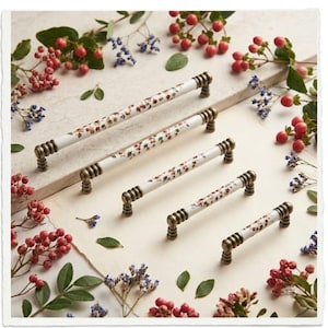 May include: Five white ceramic drawer pulls of varying lengths, each featuring a delicate floral pattern and antique brass-toned metal ends with ribbed detailing. The handles are arranged in a diagonal line, showcasing their cylindrical shape.