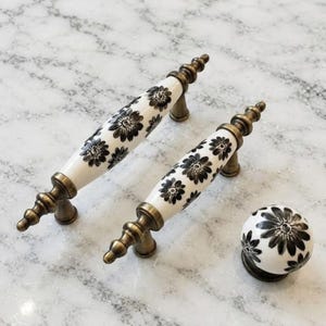 May include: Two elongated cabinet pulls and one round cabinet knob, each featuring a white ceramic surface decorated with a black floral pattern and finished with antique brass metal hardware at the ends and mounting points.